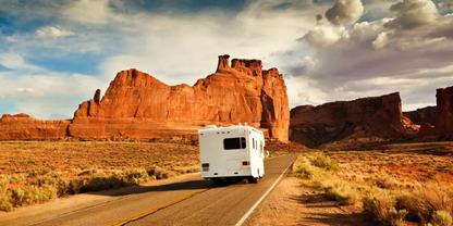 Wei&szlig;es Wohnmobil auf geteerte Zweispurstra&szlig;e, R&uuml;ckseite sichtbar, rote Felsformationen und trockenes Buschland in einer W&uuml;stenlandschaft unter blauem, teils bew&ouml;lktem Himmel