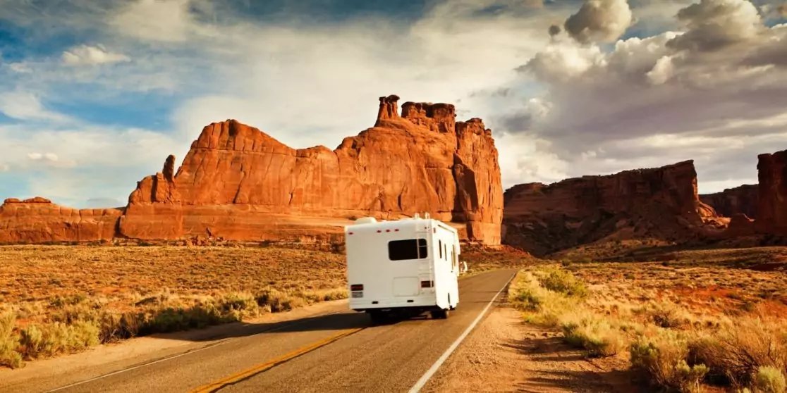 Wei&szlig;es Wohnmobil auf geteerte Zweispurstra&szlig;e, R&uuml;ckseite sichtbar, rote Felsformationen und trockenes Buschland in einer W&uuml;stenlandschaft unter blauem, teils bew&ouml;lktem Himmel