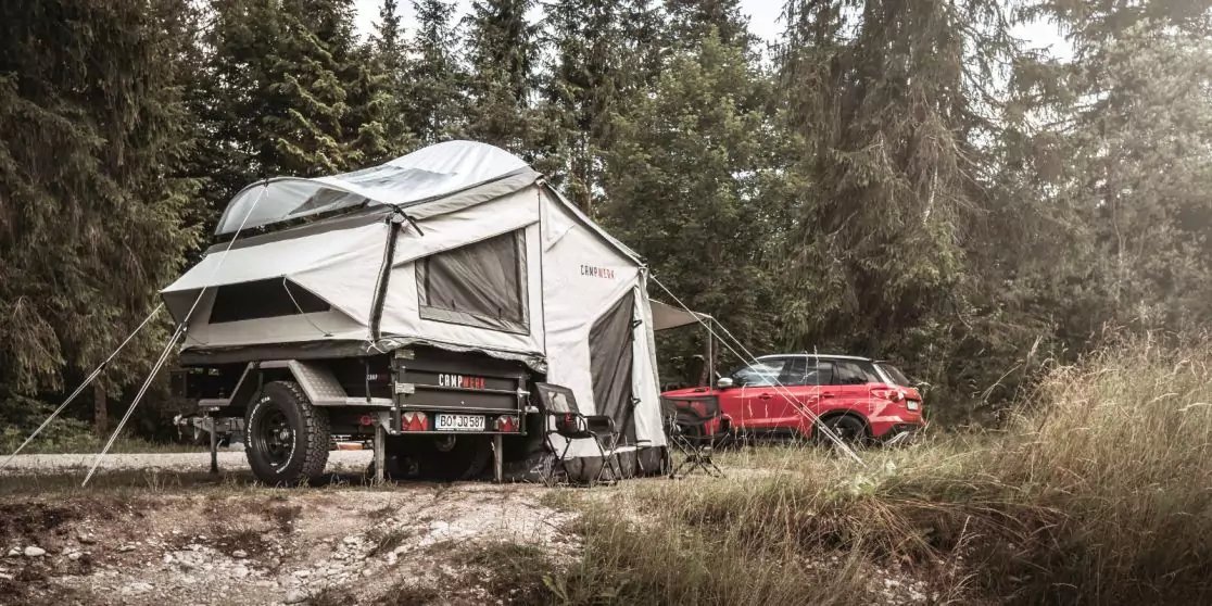 Aufgeklapptes Dachzelt auf einem Anh&auml;nger neben einem roten SUV in einer Waldlichtung mit Gras und einem unbefestigten Weg