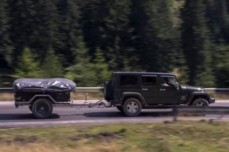 Dunkler Gel&auml;ndewagen zieht einen Anh&auml;nger mit abgedeckter Ladung auf einer Stra&szlig;e, Nadelwald im Hintergrund, Bewegungsunsch&auml;rfe im Vordergrund