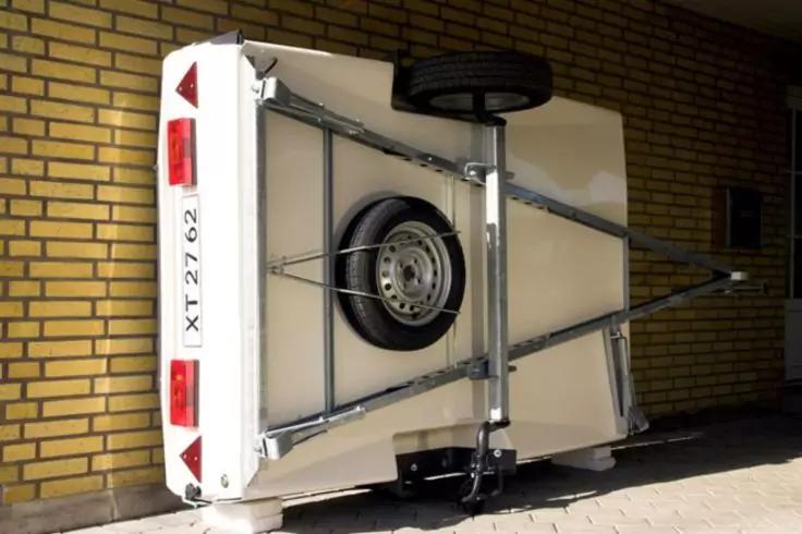 Wei&szlig;er Anh&auml;nger auf der Seite mit Reserverad, Metallrahmen, Anh&auml;ngerkupplung, gelber Backsteinwand und Gehweg