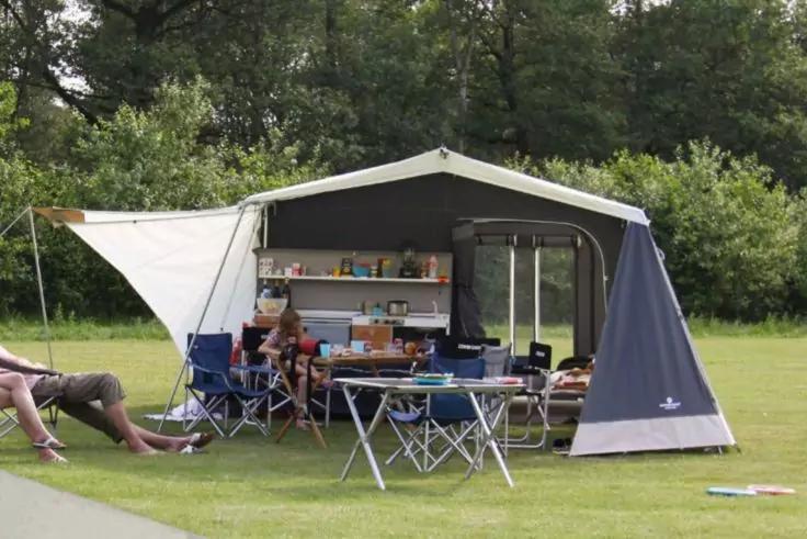 Gro&szlig;es Campingzelt mit offenem Vorzelt, Klapptisch, mehreren Campingst&uuml;hlen, Regal mit K&uuml;chenutensilien, Grasfl&auml;che und B&auml;ume im Hintergrund