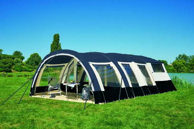 Gro&szlig;es dunkelblau-wei&szlig;es Zelt mit mehreren Fenstern und offenem Vorzelt auf gr&uuml;ner Wiese neben einem See, klarer blauer Himmel