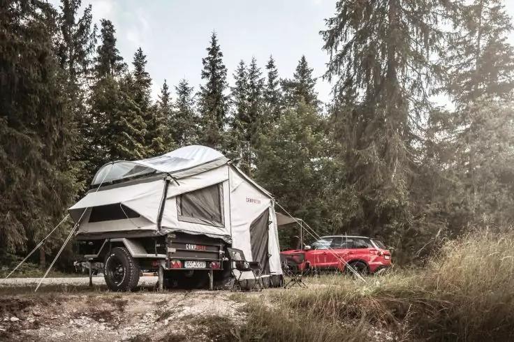 Campinganh&auml;nger mit ausgeklapptem Vorzelt neben einem roten Auto auf einem Waldweg, umgeben von Nadelb&auml;umen