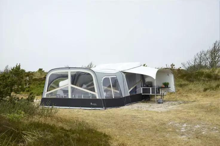 Wei&szlig;er Wohnwagen mit angebautem Vorzelt auf sandigen D&uuml;nen, kleiner Tisch und St&uuml;hle davor, D&uuml;nenvegetation und bew&ouml;lkter Himmel
