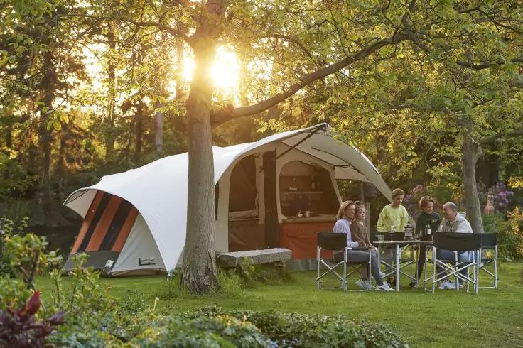 Zelt mit &uuml;berdachtem Vorbau auf einer Wiese unter B&auml;umen, Sonnenlicht f&auml;llt durch die Zweige, vier Personen sitzen an einem Campingtisch auf Klappst&uuml;hlen, Tisch mit Gegenst&auml;nden