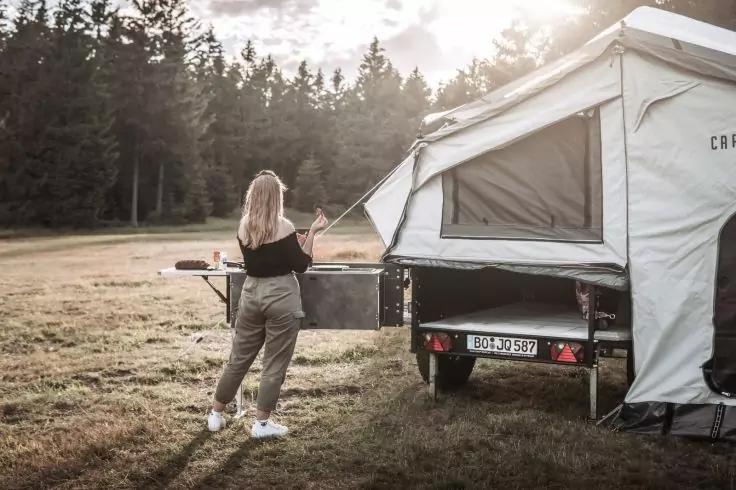 Person mit langen blonden Haaren an ausgezogenem Camping-K&uuml;chenfach eines aufgeklappten Camperanh&auml;ngers auf einer Wiese, Wald im Hintergrund