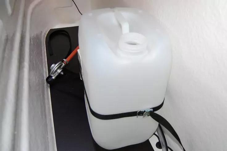 Wei&szlig;er Kunststoffkanister mit Schraubverschluss auf schwarzer Ablage, mit schwarzem Gurt befestigt; daneben ein Schraubendreher mit rotem Griff, wei&szlig;e Wand und sichtbare Rohrleitungen