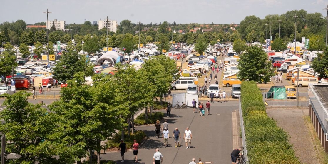 Viele Zelte und Wohnwagen auf einem gro&szlig;en Platz, B&auml;ume, ein breiter Gehweg mit Fu&szlig;g&auml;ngern, Fahrzeuge und Geb&auml;ude im Hintergrund