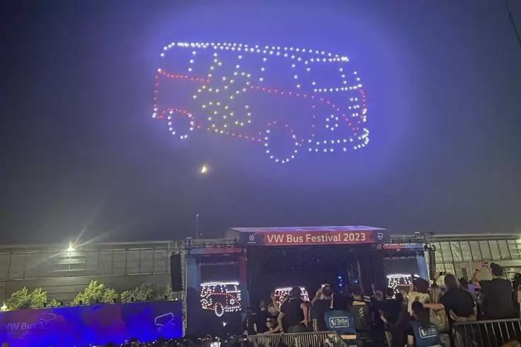 Drohnenlichter formen den Umriss eines Kleinbusses mit Blumenmotiv im Nachthimmel &uuml;ber einer B&uuml;hne mit Banner &bdquo;VW Bus Festival 2023&ldquo; und Zuschauern