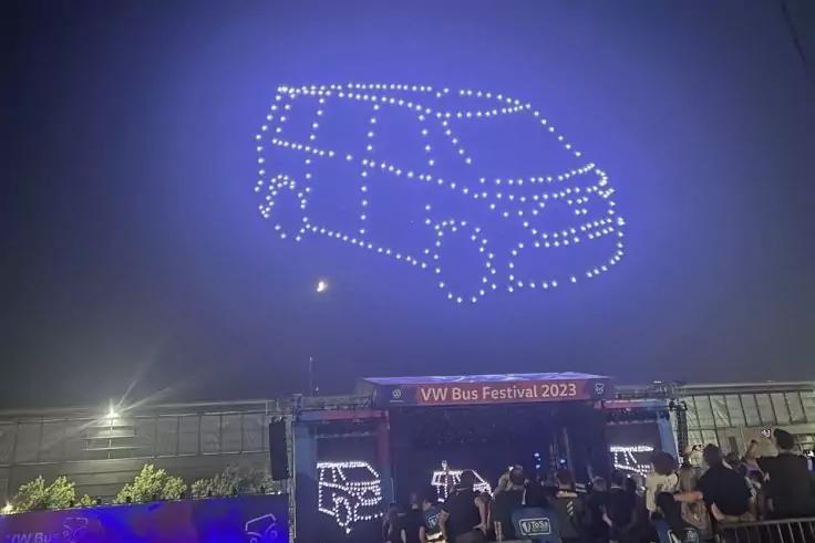 Leuchtpunkte formen im blauen Nachthimmel die Umrisse eines Vans; darunter B&uuml;hne mit Banner "VW Bus Festival 2023" und Menschen