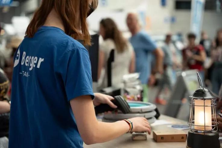 Person in blauem T‑Shirt h&auml;lt einen Handscanner &uuml;ber Waren an einer Kasse; mehrere unscharfe Personen im Hintergrund