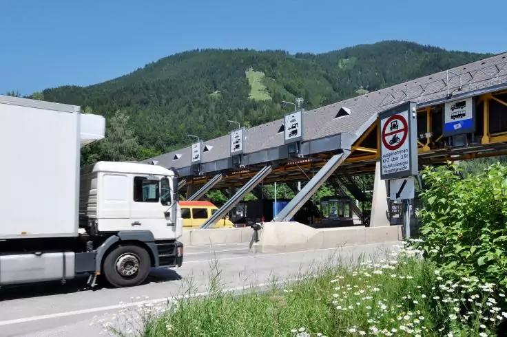 Wei&szlig;er Lkw links auf der Stra&szlig;e, dahinter ein gelbes Fahrzeug. Rechts eine erh&ouml;hte, &uuml;berdachte Holzstruktur mit schr&auml;gen Glasg&auml;ngen. Verbotsschild an einem Pfosten, Wiese mit Blumen im Vordergrund, bewaldeter H&uuml;gel und blauer Himmel im Hintergrund
