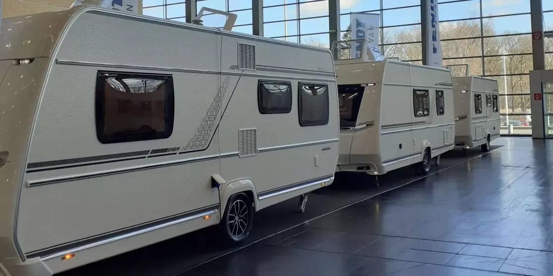 Drei hellbeige Wohnwagen nebeneinander in einer hellen Halle vor gro&szlig;en Glasfenstern