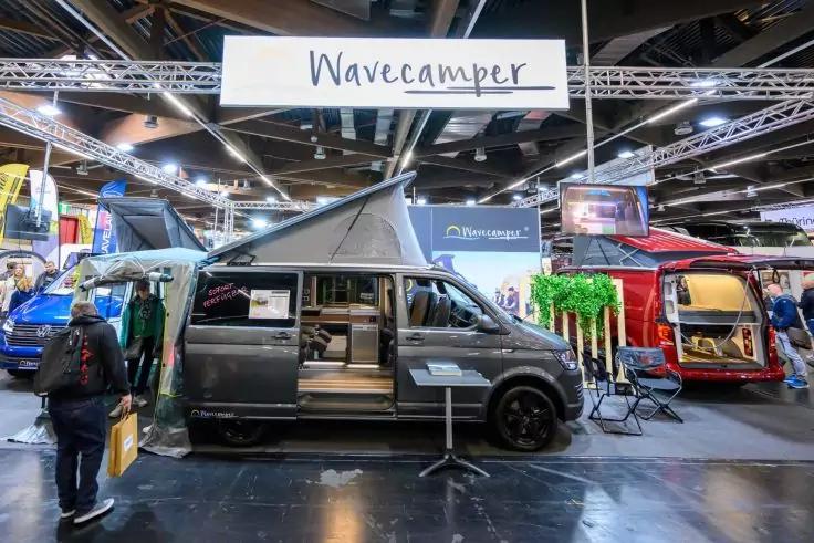 Dunkelgrauer Campervan mit aufgeklapptem Hochdach und offener Seitent&uuml;r unter Schild 'Wavecamper' in einer Halle; weitere bunte Vans und eine Person mit Rucksack im Hintergrund