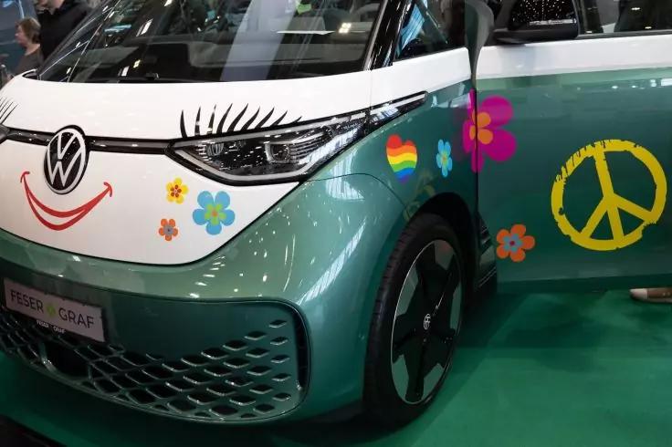 Vorderer linker Bereich eines t&uuml;rkisfarbenen Volkswagen mit wei&szlig;er Motorhaube, VW‑Logo, Wimpernaufkleber am Scheinwerfer, Smiley-Aufkleber unter dem Logo, Blumen- und Regenbogenherz-Aufkleber an der T&uuml;r, gro&szlig;es gelbes Peace‑Symbol, sichtbares Vorderr