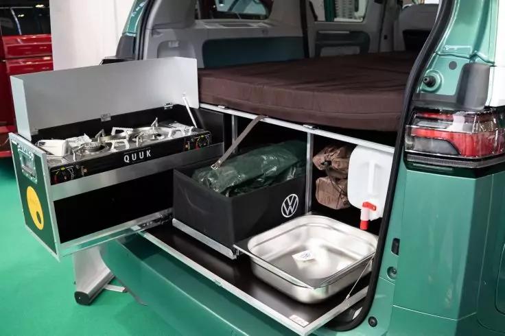 Ge&ouml;ffnetes Heck eines gr&uuml;nen Vans mit ausziehbarer Metalllade; Edelstahlsp&uuml;le, schwarzer Beh&auml;lter mit Volkswagen-Logo, gr&uuml;ner Stoffbeutel, wei&szlig;e Plastikflasche mit rotem Verschluss und gefaltete braune Polsterfl&auml;che