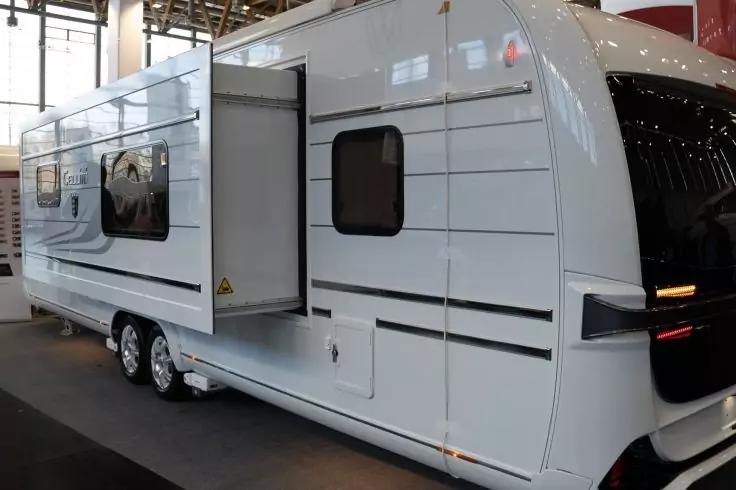 Wei&szlig;er Wohnwagen mit ausgefahrenem Seitenmodul, zwei Achsen (vier R&auml;der), mehreren rechteckigen Fenstern, schwarzen horizontalen Streifen und abgerundeter Front mit dunklem Frontfenster; Hintergrund mit gro&szlig;en Fenstern und Deckenstreben