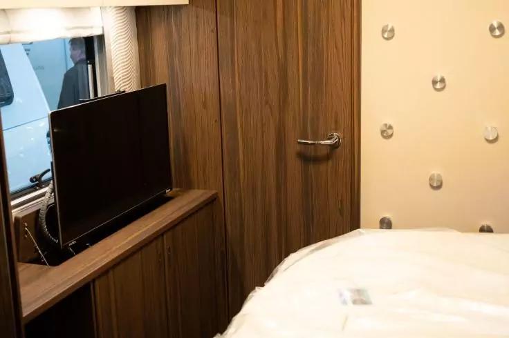 Fernseher auf niedrigem Holzschrank, neben einer holzvert&auml;felten Wand mit geschlossener T&uuml;r und Metallgriff; rechts ein Bett mit wei&szlig;er Bettdecke; links ein Fenster mit Vorhang