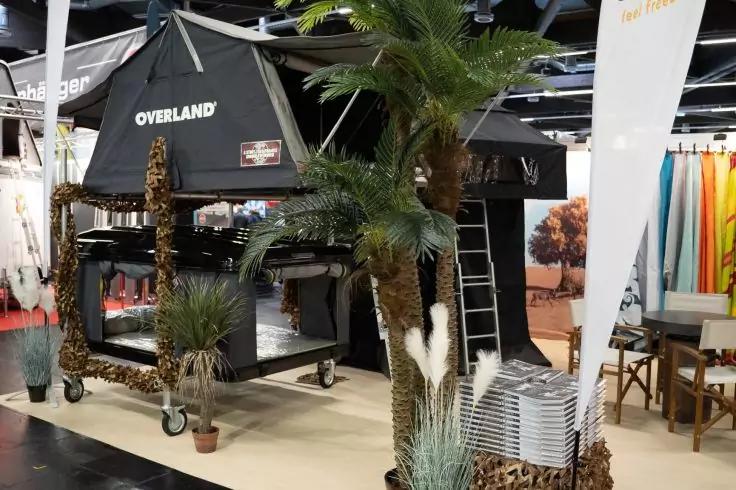 Schwarzes Dachzelt mit Aufschrift 'OVERLAND' auf einem fahrbaren Gestell, angelehnte Leiter, mehrere dekorative Palmen und Pampasgras, gestapelte silberne Tabletts, Banner sowie Tische mit St&uuml;hlen im Hintergrund