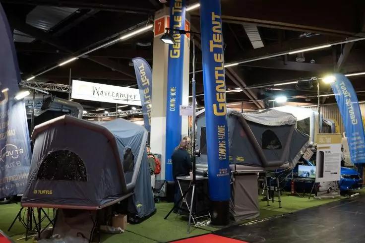 Hallenraum mit mehreren ausgestellten Zelten, blauen S&auml;ulen und Fahnen mit der Aufschrift &sbquo;GENTLE TENT&lsquo;, Tische und ein Monitor, eine Person steht am Stand