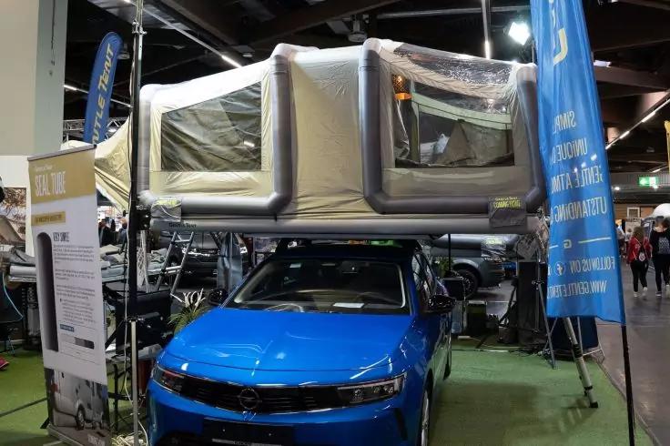 Blauer Pkw mit aufgeklapptem Dachzelt &uuml;ber dem Fahrzeug, in einer Messehalle mit Bannern im Hintergrund