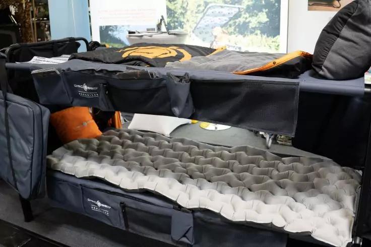 Zwei &uuml;bereinander angeordnete Campingbetten mit grauen aufblasbaren Matratzen, einem orangefarbenen Kissen, schwarzen Stoffrahmen mit Logos und einem Fenster mit B&auml;umen im Hintergrund