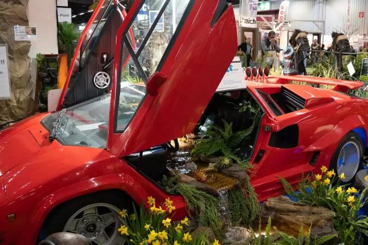 Rotes Sportauto mit ge&ouml;ffneten Scherent&uuml;ren; Innenraum und Motorraum mit Pflanzen und Moos gef&uuml;llt; gelbe Blumen und Holzst&uuml;cke im Vordergrund; Personen im Hintergrund