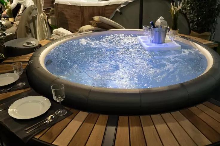 Runder Whirlpool mit blau beleuchtetem, sprudelndem Wasser auf Holzdeck; am Rand Tisch mit Teller, Glas und Besteck; Pflanzen und teilweise sichtbare Person im Hintergrund