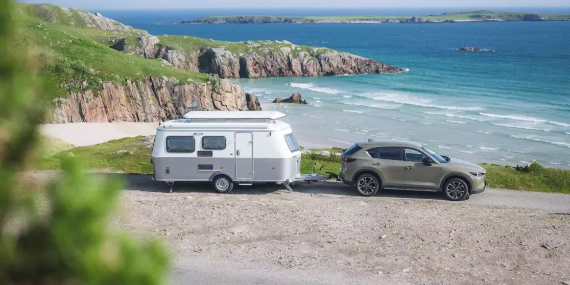 Auto mit wei&szlig;em Wohnwagen auf einer K&uuml;stenstra&szlig;e neben Sandstrand und felsigen Klippen, blaues Meer und Insel im Hintergrund, Gras im Vordergrund