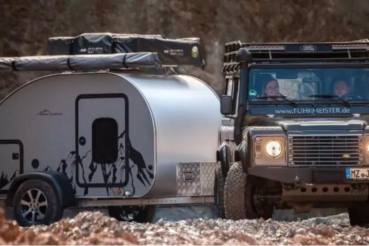 Dunkelgr&uuml;nes Fahrzeug mit Dachgep&auml;cktr&auml;ger, zwei Personen im Fahrerhaus; dahinter ein silberner, abgerundeter Anh&auml;nger mit schwarzen Personen‑Silhouetten auf der Seitenwand; staubiger, felsiger Untergrund