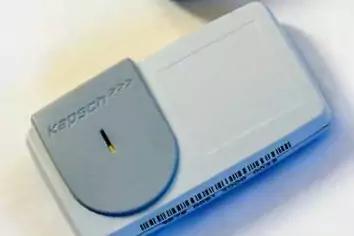 Wei&szlig;es rechteckiges Ger&auml;t auf hellem Untergrund, links runde graue Abdeckung mit schmalem dunklem Schlitz, Barcode-Etikett am unteren Rand