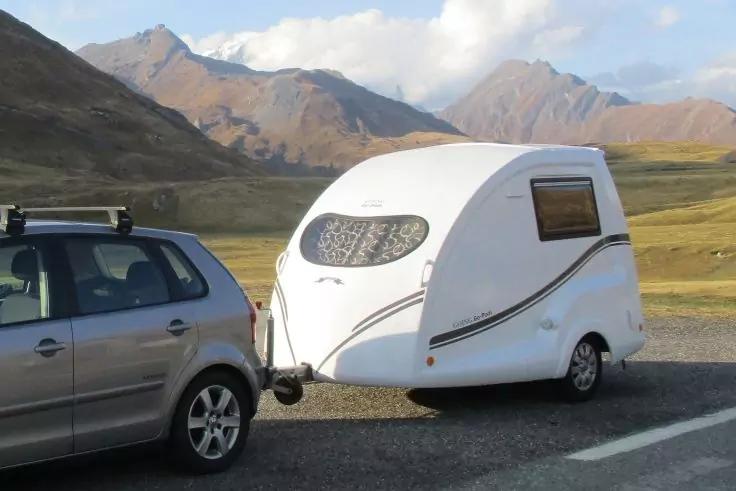 Silbernes Auto zieht einen kleinen wei&szlig;en Wohnanh&auml;nger auf einer Stra&szlig;e, grasige Ebene und Berge im Hintergrund, teils bew&ouml;lkter Himmel