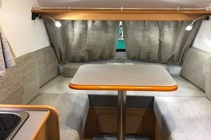 Tisch in der Mitte, zwei gegen&uuml;berliegende gepolsterte Sitzb&auml;nke mit Kissen, Vorh&auml;nge vor einem Fenster, zwei Leseleuchten, rechteckige Tischplatte mit Holzkante, Sp&uuml;le am linken Bildrand