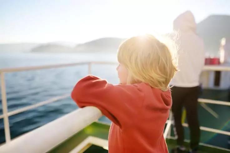 Kind mit blondem Haar in rotem Pullover lehnt am Reling eines Schiffes, Blick aufs Meer, Sonnenreflexe und entfernte H&uuml;gel; im Hintergrund Person in wei&szlig;er Jacke
