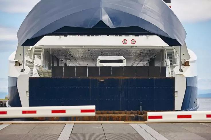 Frontansicht einer wei&szlig;en und blauen F&auml;hre mit sichtbarer zentraler Fahrzeug&ouml;ffnung, im Vordergrund eine rot-wei&szlig;e Schranke und der Hafenboden