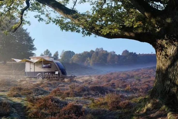 Kleiner Wohnanh&auml;nger mit ausgefahrener Markise auf nebelverhangener Heide, im Vordergrund ein gro&szlig;er Baumstamm mit &Auml;sten, im Hintergrund eine Baumreihe