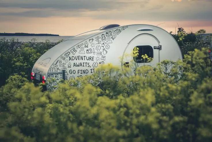 Kleiner wei&szlig;er Teardrop-Camper mit Illustrationen und Schrift 'ADVENTURE AWAITS, GO FIND IT', umgeben von gelben Wildblumen, dahinter Wasser und Abendhimmel