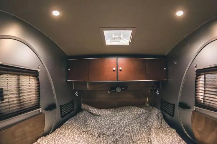 Bett mit gemusterter Decke, dar&uuml;ber h&auml;ngende Schr&auml;nke, zwei seitliche Fenster mit Jalousien, gew&ouml;lbte W&auml;nde und Deckenlicht