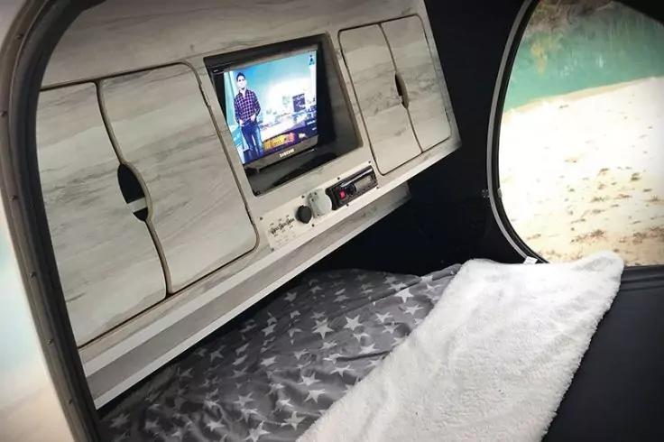 Liegefl&auml;che mit sternenbedruckter Decke und wei&szlig;em Handtuch, dar&uuml;ber Holzschrank mit kleinem Bildschirm, rundes Fenster mit Blick aufs Wasser