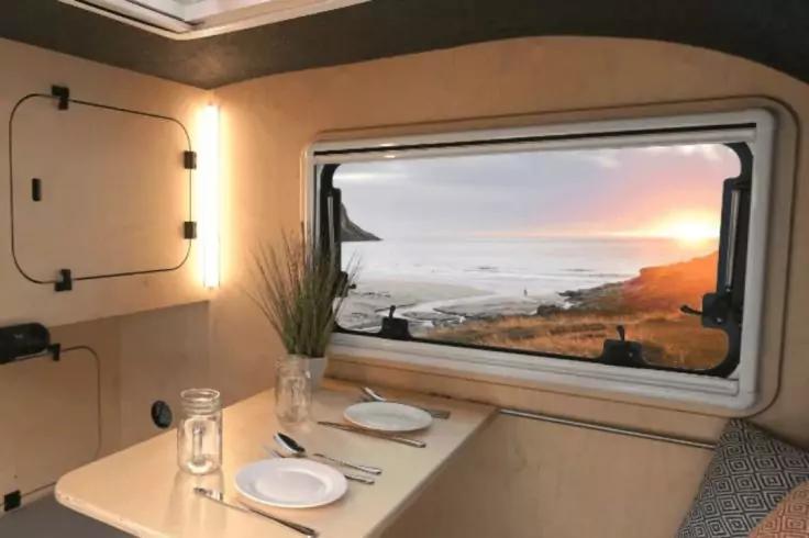 Innenraum eines Wohnmobils, Tisch f&uuml;r zwei mit Tellern, Besteck, Glas und kleiner Pflanze; Blick durch gro&szlig;es Fenster auf K&uuml;ste mit Meer, Felsen und Sonne am Horizont