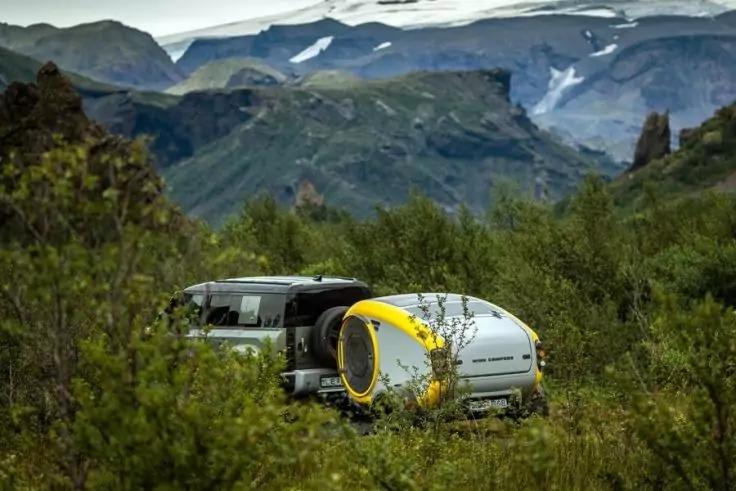 Wei&szlig;-gelber Teardrop-Camper hinter einem grauen Fahrzeug in gr&uuml;nen B&uuml;schen, Berge mit Schneefeldern im Hintergrund