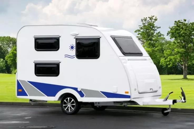 Wei&szlig;er Wohnwagen mit blauen Streifen, drei Seitenfenstern, vorderem Dachfenster, einer Achse mit Rad und Anh&auml;ngerkupplung, auf Asphalt vor Grasfl&auml;che und B&auml;umen unter bew&ouml;lktem Himmel