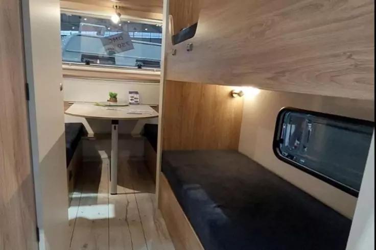 Schmaler, holzverkleideter Innenraum mit Sitzbank und Tisch am Ende, kleiner Topf mit Pflanze auf dem Tisch, rechte Seite Liegefl&auml;che mit dunklem Polster, rechteckiges Fenster, Decken- und Wandbeleuchtung