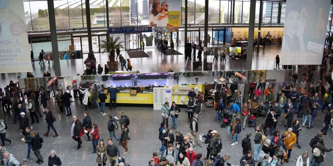 Gro&szlig;e helle Halle mit vielen Menschen, zentraler gelber Schalter, St&auml;nde, gro&szlig;e Fensterfront und h&auml;ngende Werbebanner