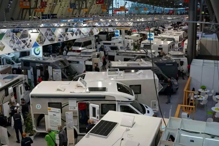 Mehrere weiße Wohnmobile in einer großen Halle mit Metallträgern, Personen zwischen den Fahrzeugen, Informationsschildern und einem Wohnmobil mit Solarpanel auf dem Dach