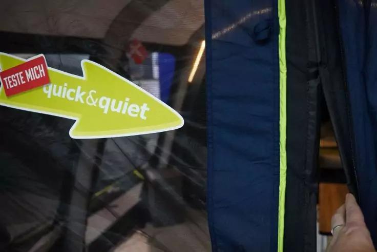 Grüner Pfeil-Aufkleber mit weißer Schrift 'quick&quiet' und rotem Schild 'TESTE MICH'; daneben dunkelblauer Stoff mit neongrünem Reißverschluss, eine Hand hält den Stoff; im Hintergrund ein Rad mit Speichen