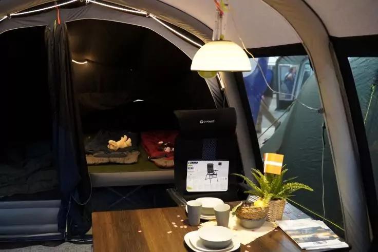 Innenraum eines Zeltes mit einem Holztisch, auf dem Teller, Tassen, eine Topfpflanze und eine Lampe stehen; rechts ein Stuhl mit Preisschild; hinten ein Schlafbereich mit Kissen und Decken