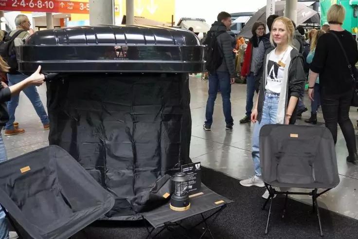 Aufnahme in einer Halle mit einem offenen großen schwarzen Koffer, zwei schwarzen Taschen auf Ständern, einer kleinen Lampe auf einem Tisch und mehreren Personen im Hintergrund