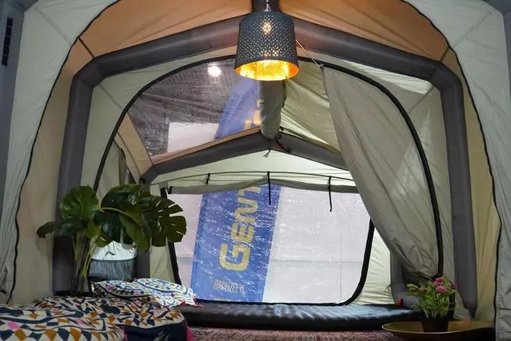 Innenansicht eines Zelts mit aufgehängter Lampe, offenem Vordereingang, Matratze mit gemustertem Überwurf, Topfpflanze links, kleine Topfpflanze rechts, durch das Fenster sichtbar eine blaue Fahne mit weißer Schrift